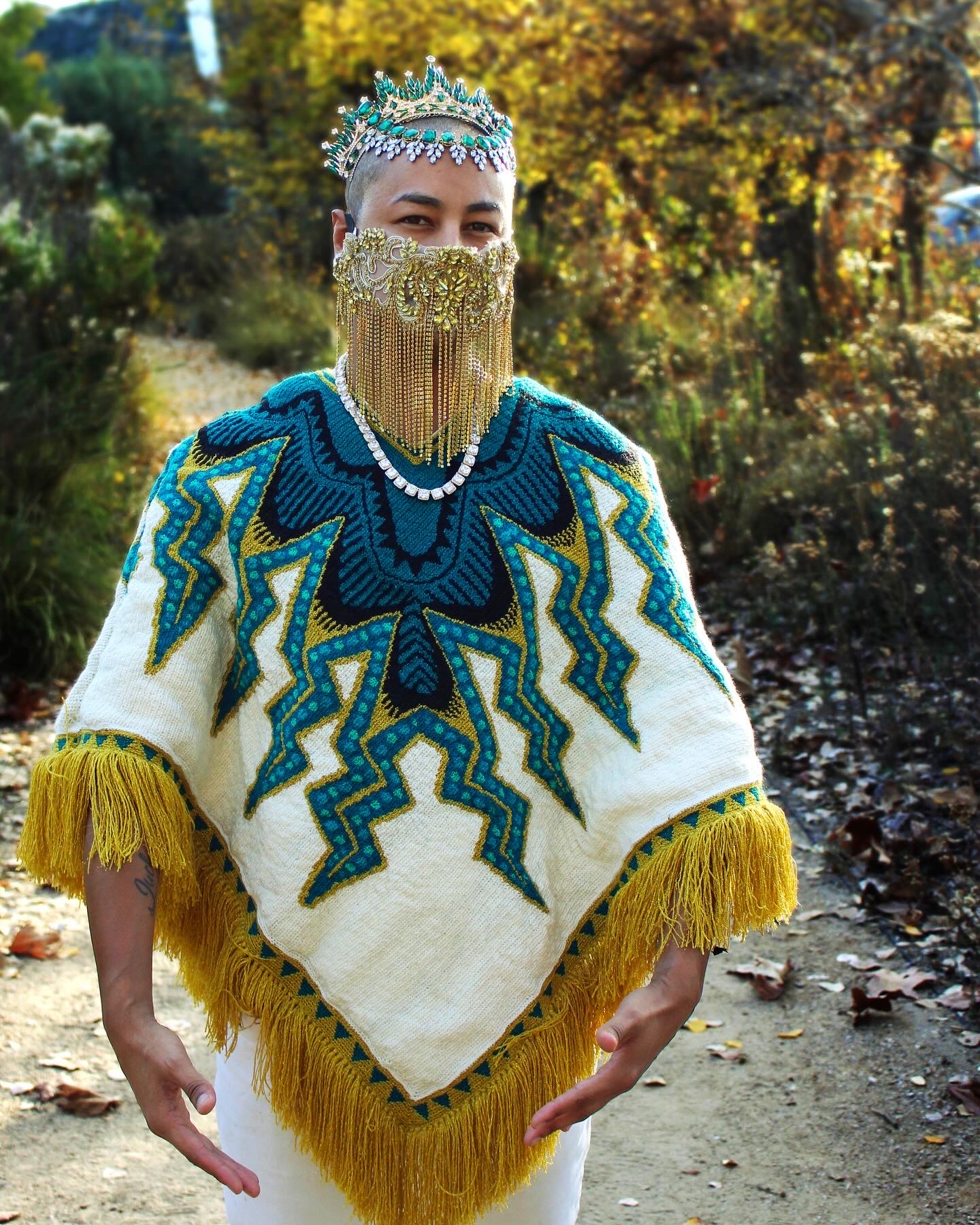 Person wearing a beaded crown and a decorative, fringed garment outdoors with trees and fallen leaves.