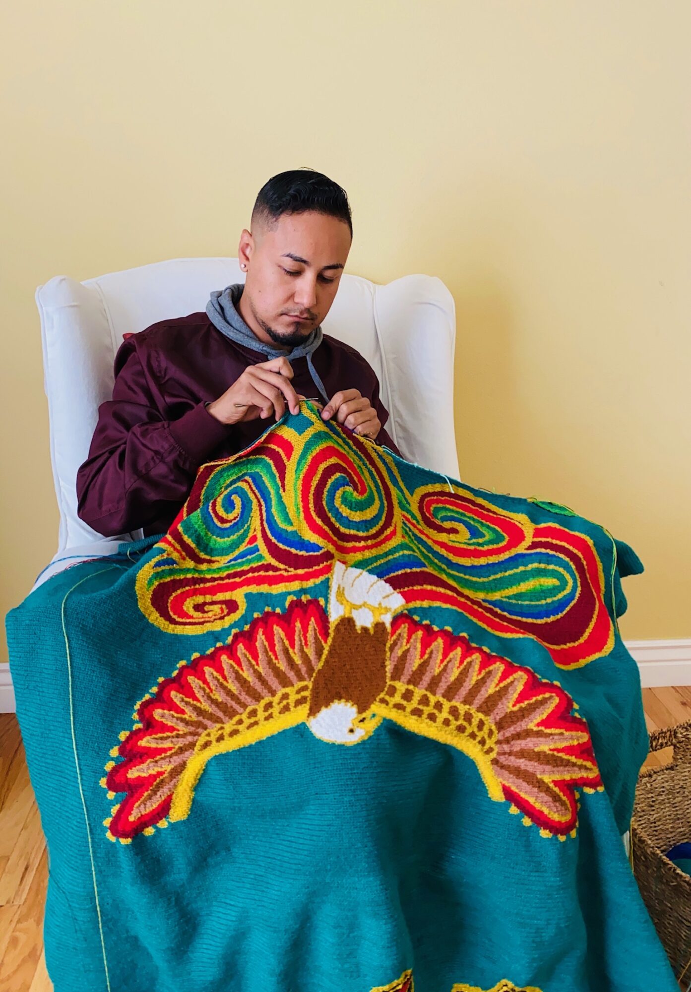 Person sitting in a white armchair, knitting a colorful blanket with a large eagle design, against a yellow wall.