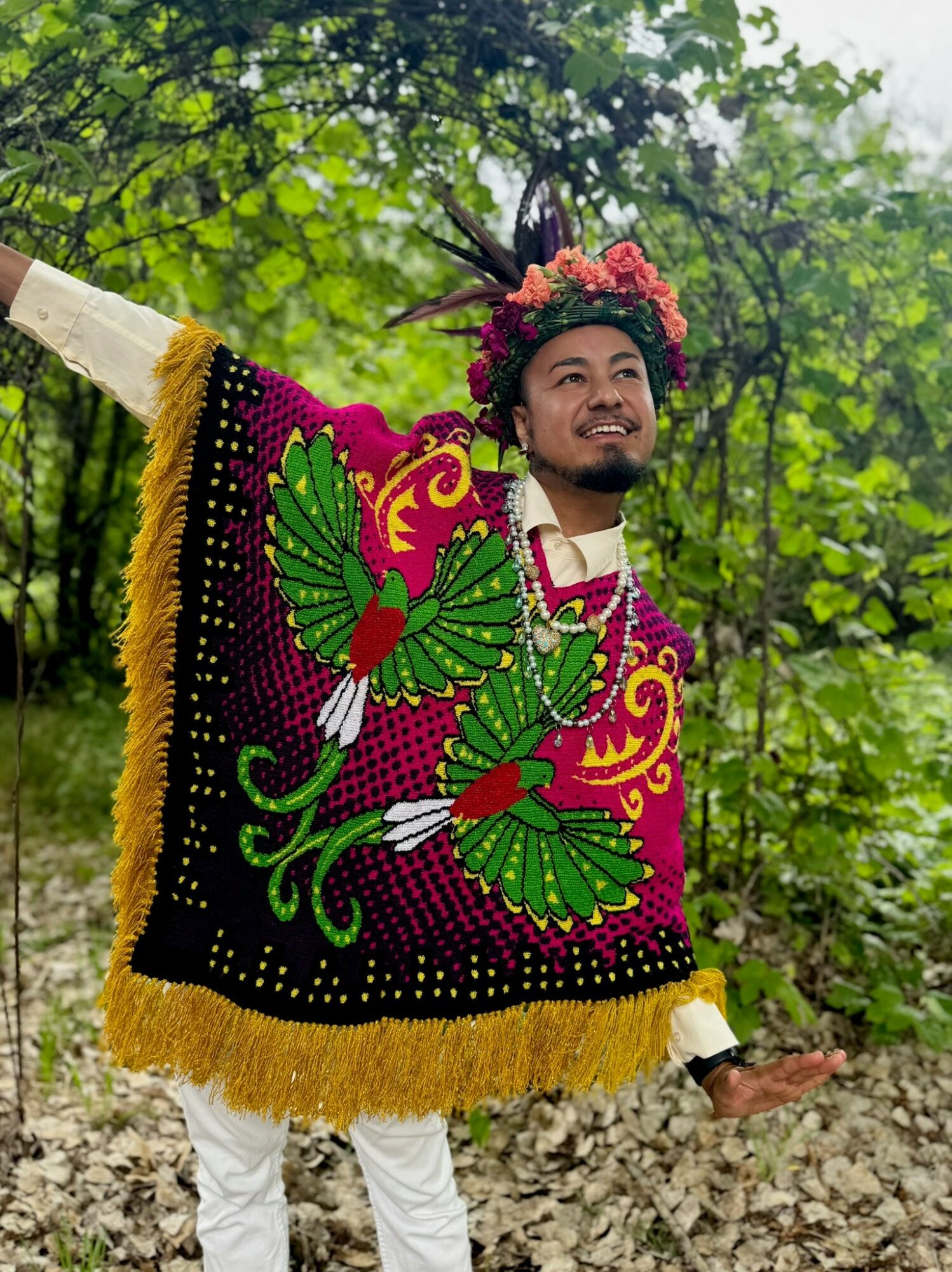 Person wearing a colorful, embroidered traditional garment with a floral headpiece, standing outdoors in a forested area.