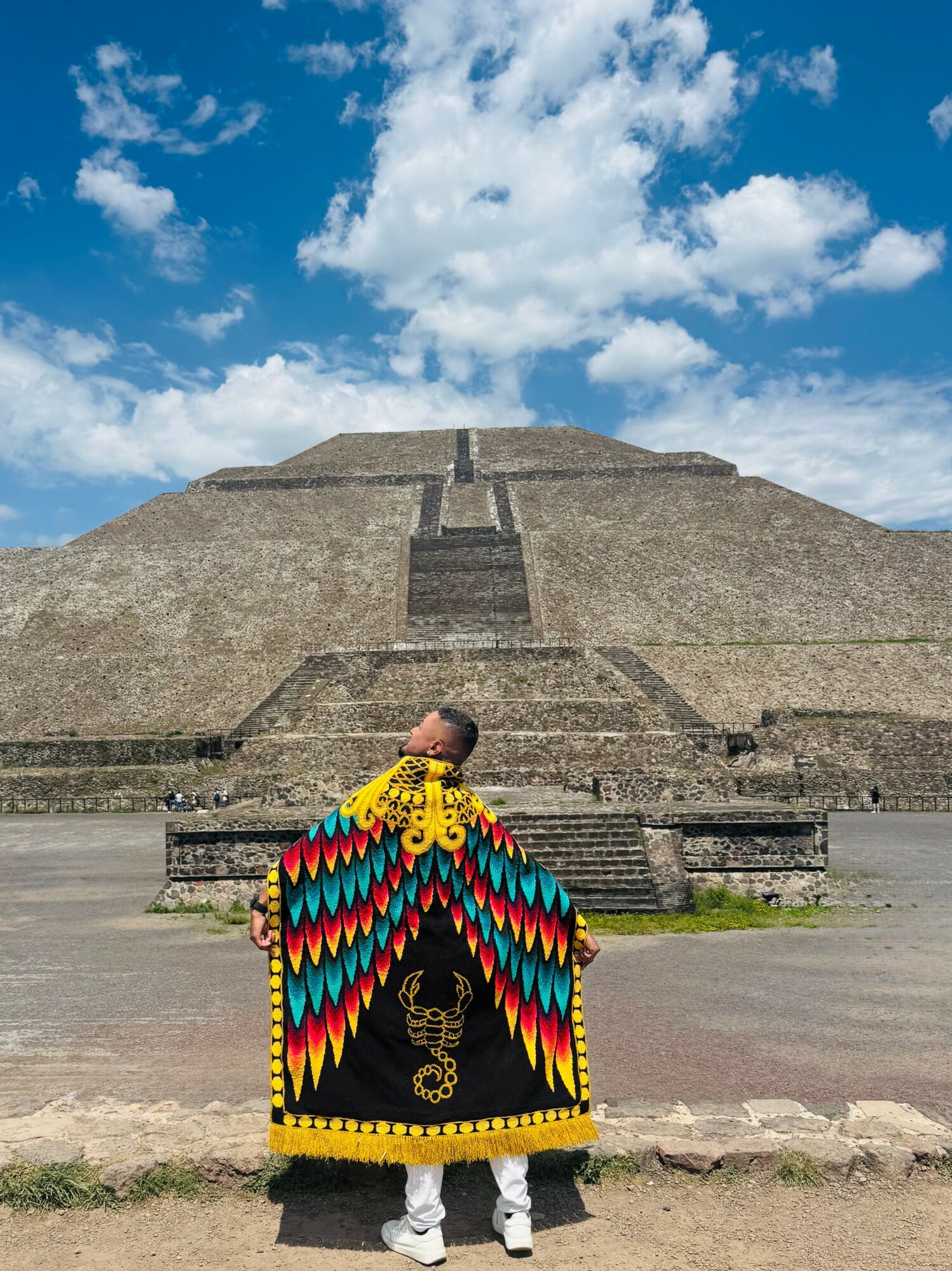 Person wearing colorful traditional clothing standing in front of a large stepped pyramid under a blue sky with clouds.