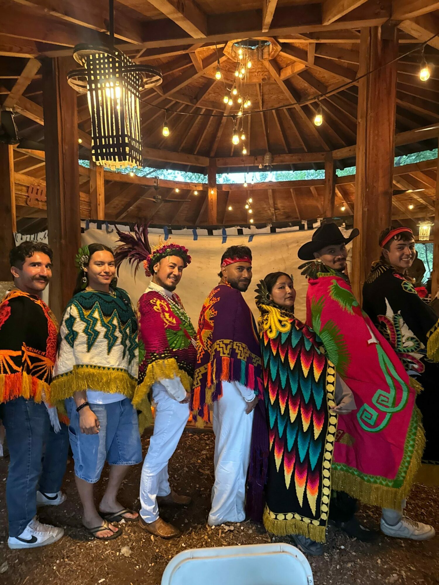 Group of people in colorful traditional clothing standing indoors with wooden beams and hanging lights.