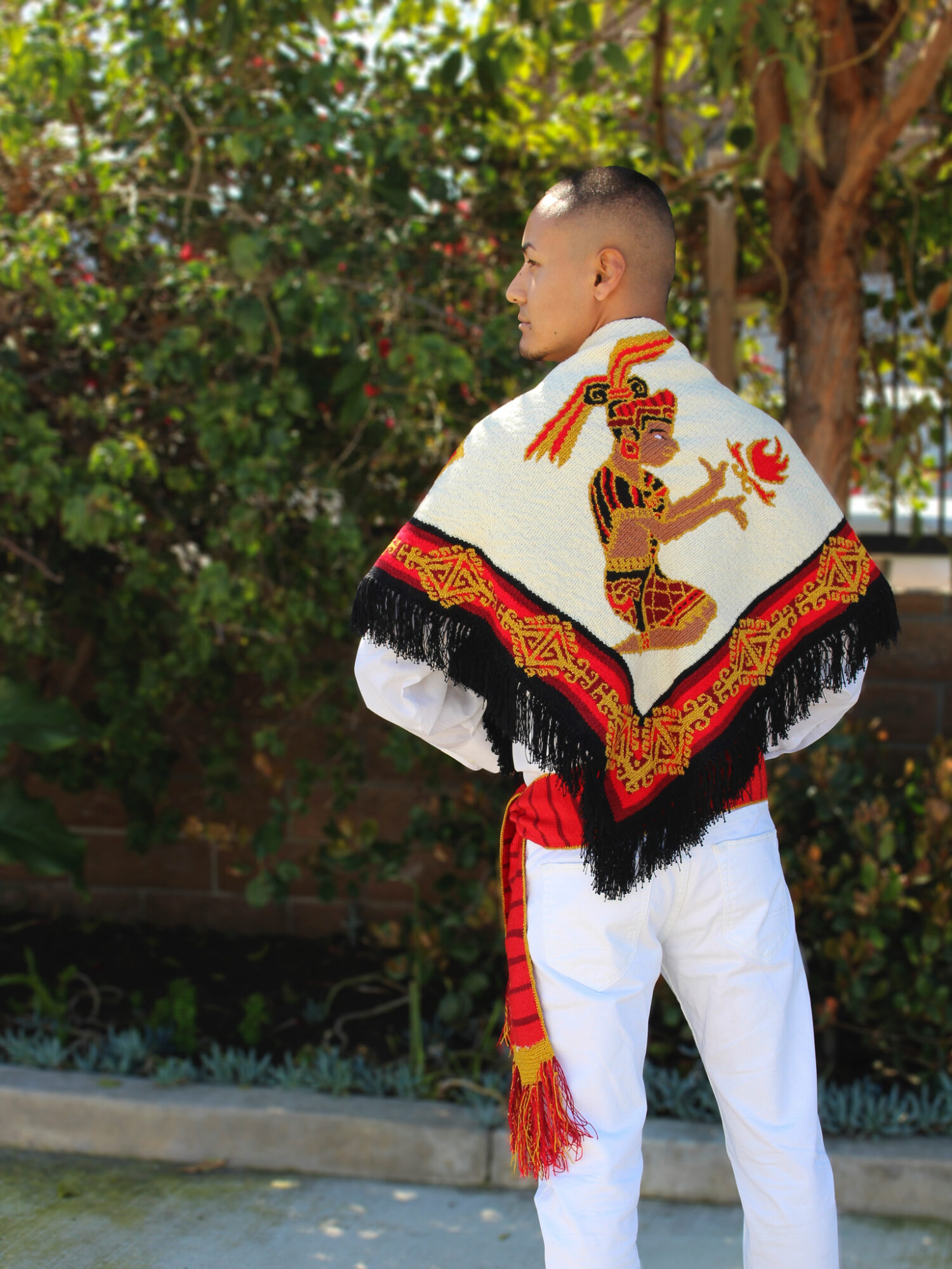 Young man in traditional attire with a colorful shawl featuring a deity, outdoors with trees and greenery.