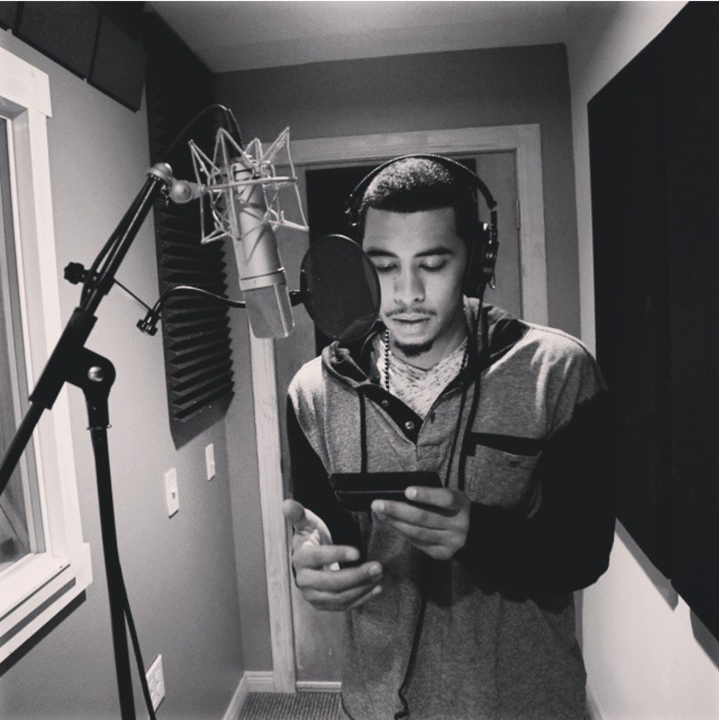 Young man in a recording studio, wearing headphones, looking at a smartphone, standing in front of a microphone, with soundproofing panels.