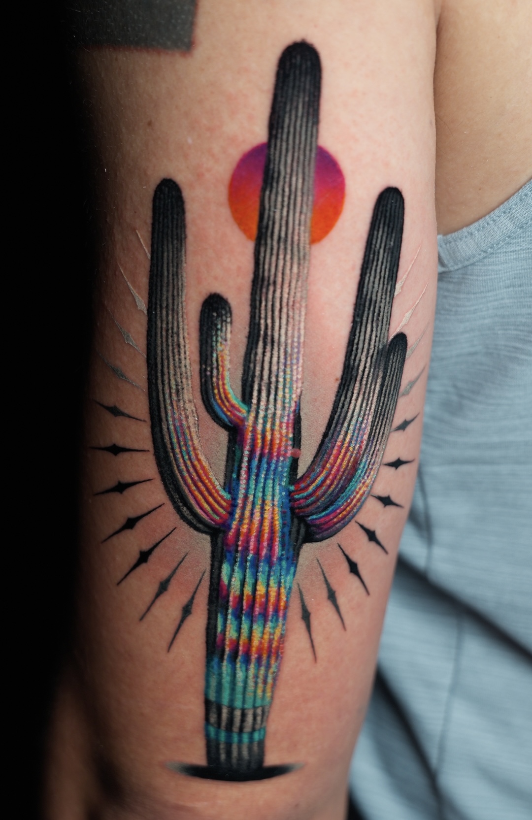 Colorful cactus tattoo with a red sun behind it on person's arm, with black lines radiating outward.