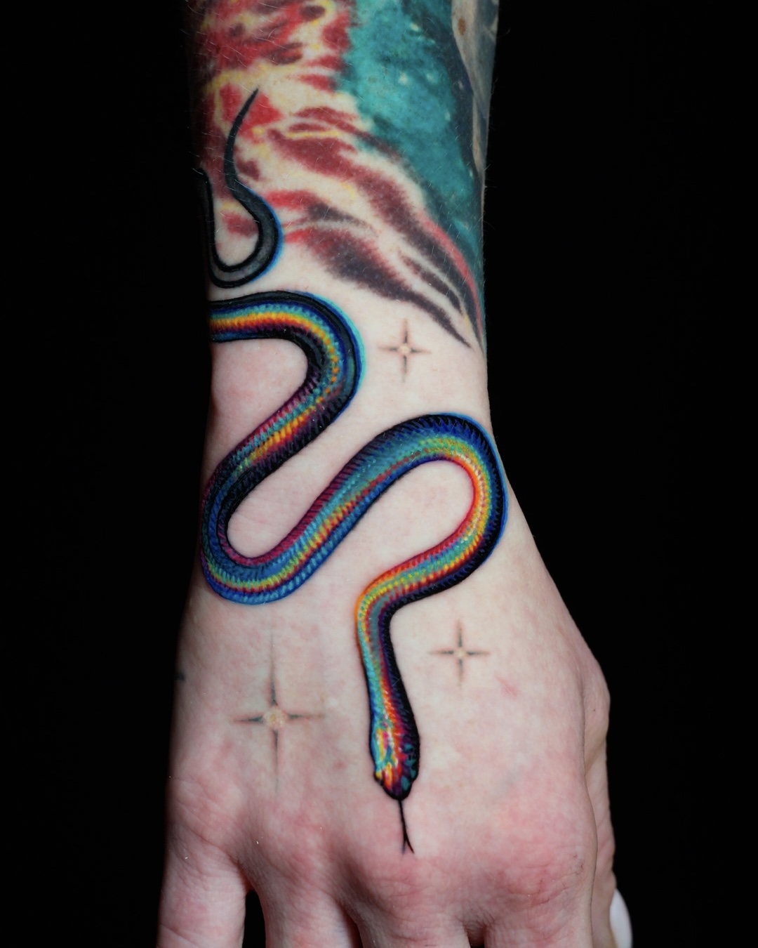 Colorful snake tattoo on person's hand and wrist with stars and abstract designs, against a black background.