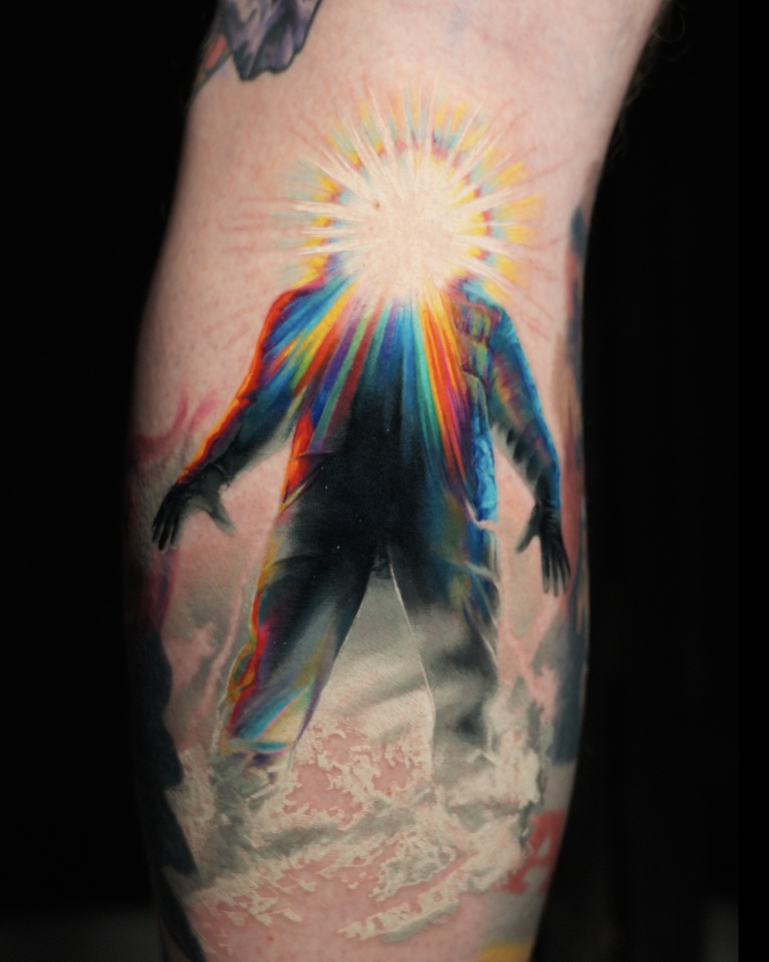 Tattoo of a person with a rainbow-colored light emanating from their head, standing on clouds, with a dark background.