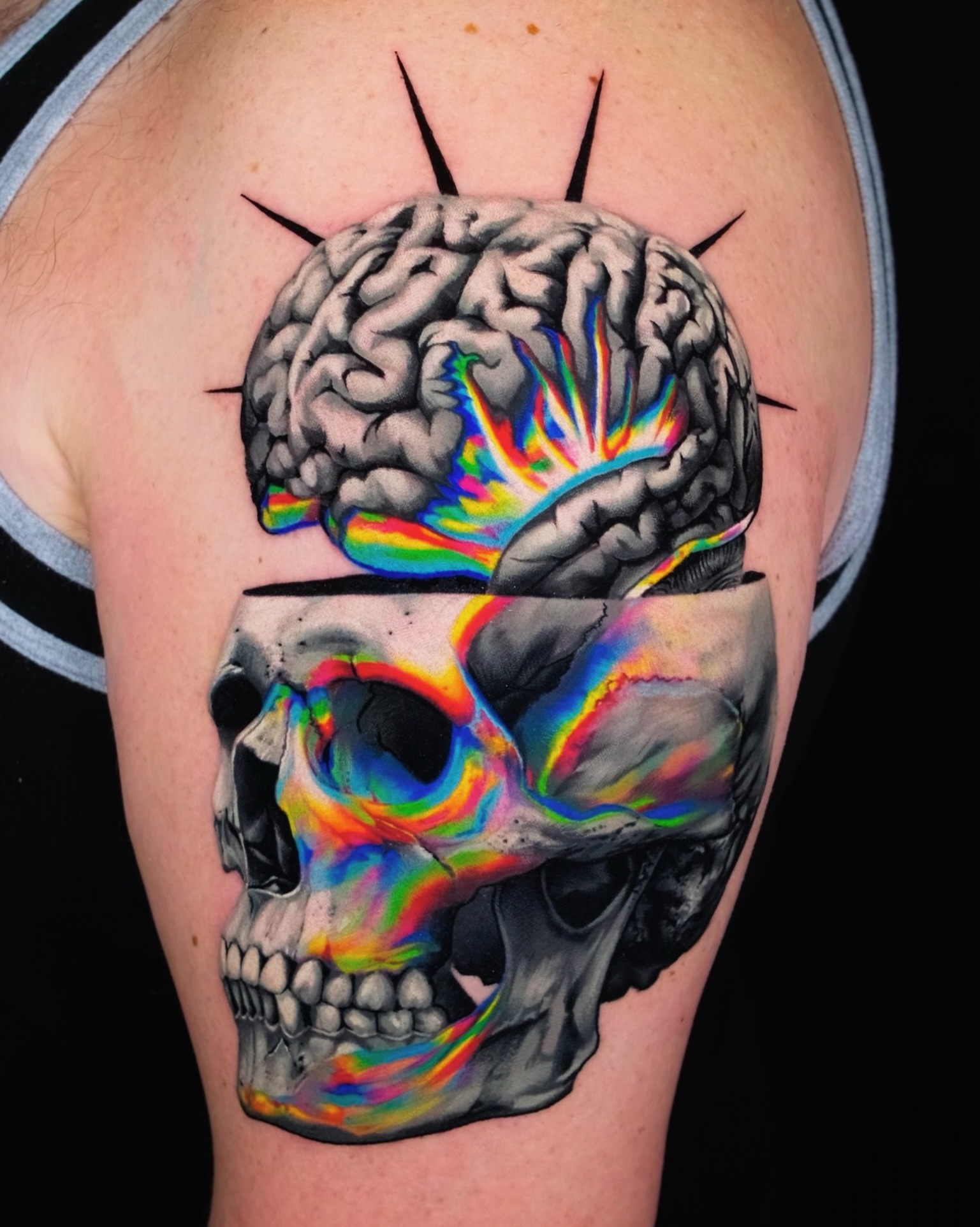 Tattoo of a brain above a colorful skull on upper arm, with black lines radiating outward, against a dark background.