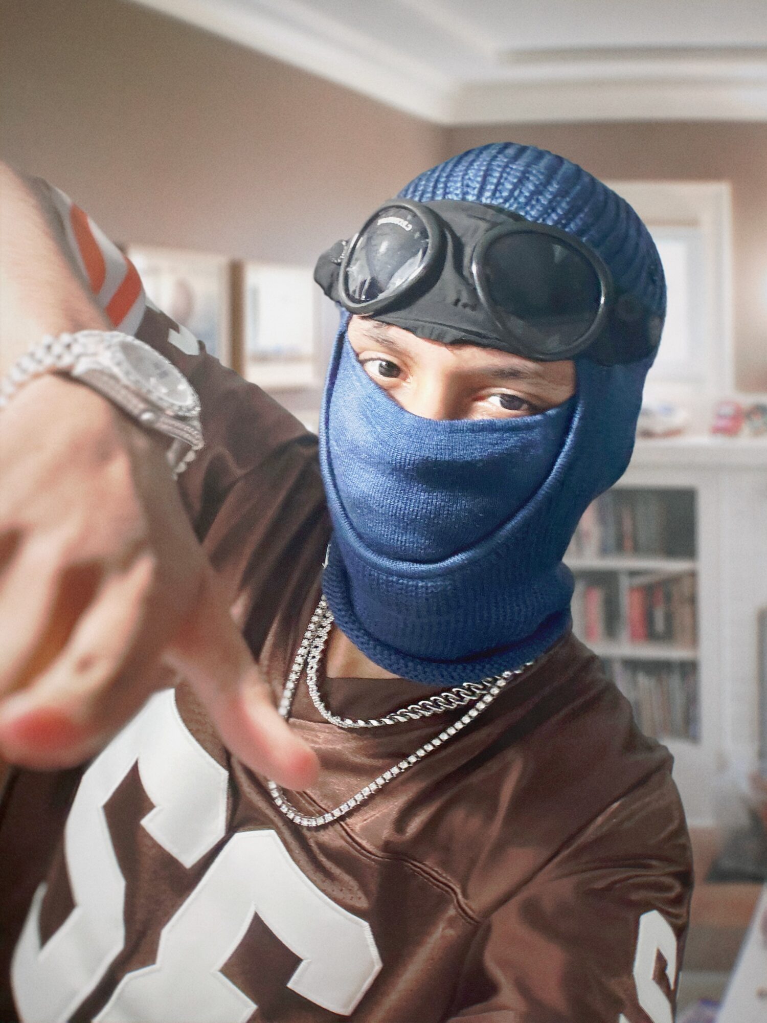Person wearing a blue ski mask, goggles, and jewelry, pointing at the camera indoors.