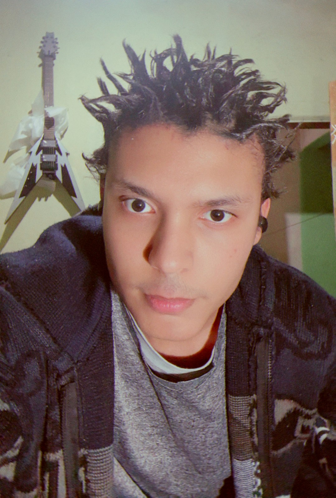 Young person with spiky hair, wearing a gray shirt and dark jacket, looking at the camera indoors.