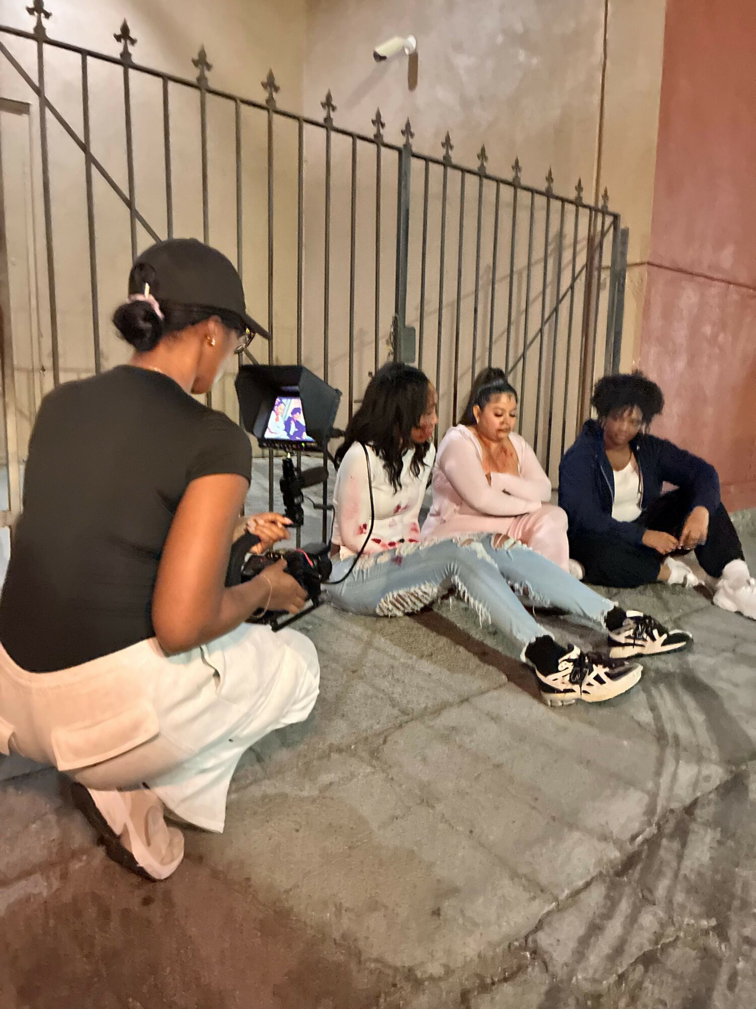 Four people sitting on the floor, two with laptops, in a room with a metal railing and a pink wall.