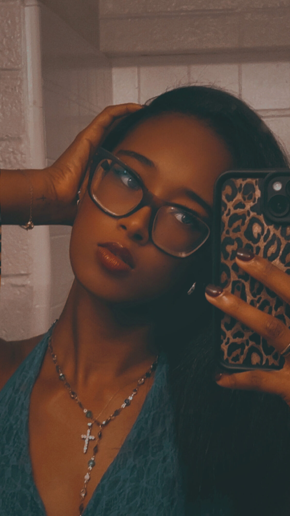 Young woman with glasses and dark hair taking a mirror selfie, wearing a necklace with a cross, in a room with white brick wall.