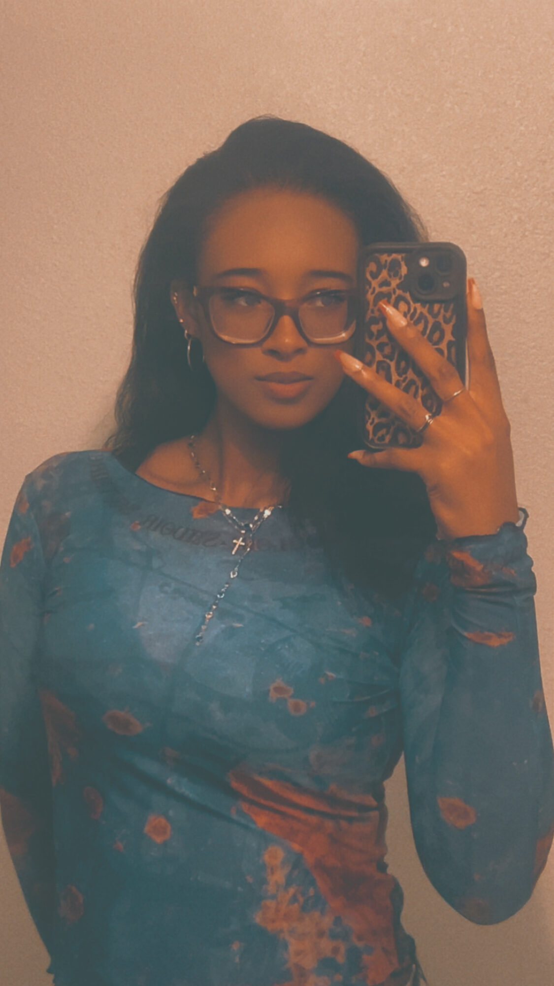 Young woman with glasses and long dark hair taking a mirror selfie, wearing a blue patterned top.