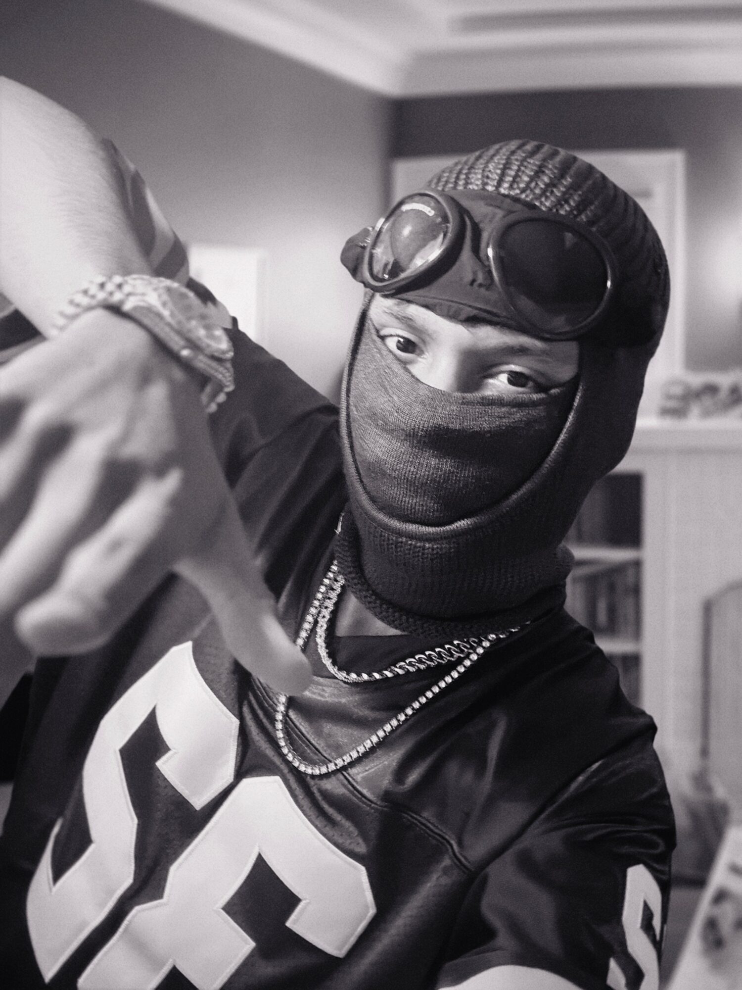 Person wearing goggles, mask, and hoodie, making a hand gesture, indoors with a bookshelf in the background.