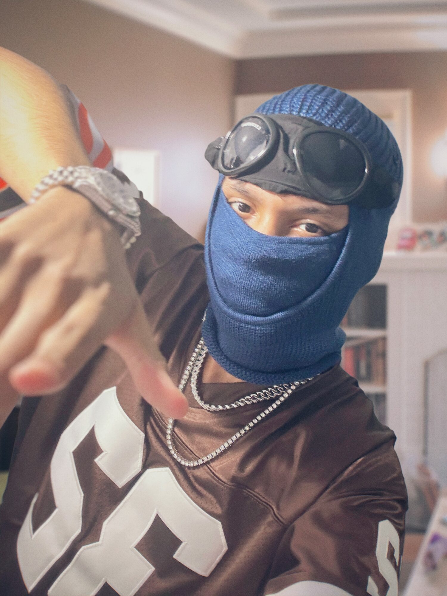 Person wearing a blue ski mask, goggles, and a beanie, making a gesture with their hand in an indoor setting.