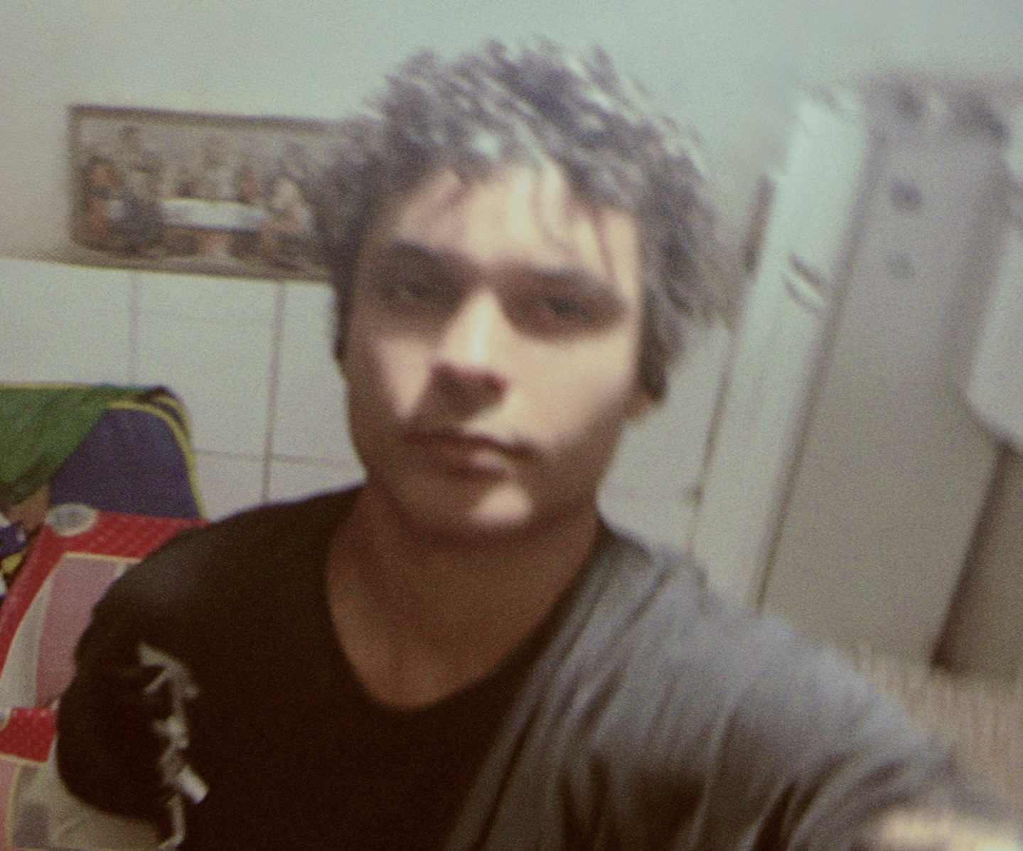 Young man with curly hair taking a selfie in a room with tiled walls and a refrigerator.