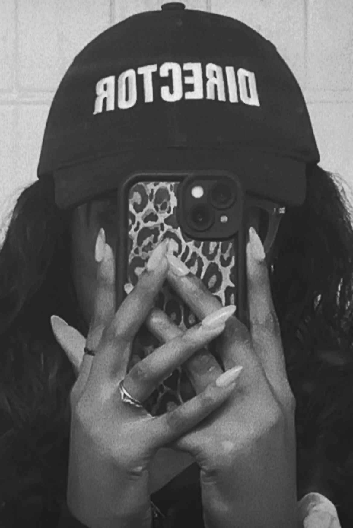 Person wearing a cap with 'DIRECTOR' text, taking a mirror selfie with a phone, black and white image.