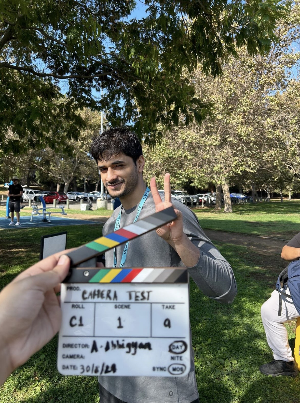 Young man outdoors holding a clapperboard, smiling, trees and people in background, sunny day.
