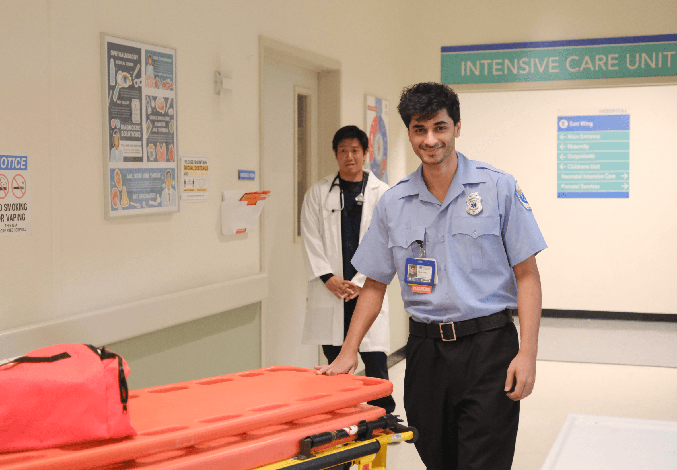 Two healthcare professionals in a hospital corridor, one smiling and pushing a stretcher, the other standing behind him. Sign reads 'Intensive Care Unit'.
