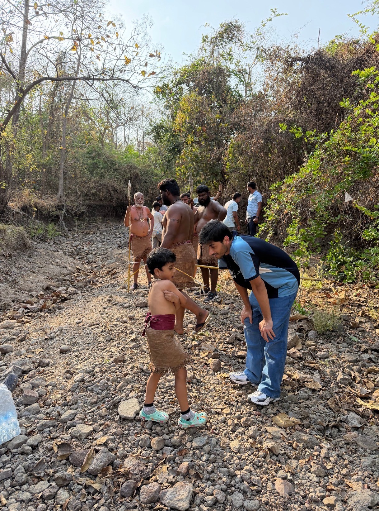 Group of people, including children, walking on rocky terrain with trees and bushes in the background.
