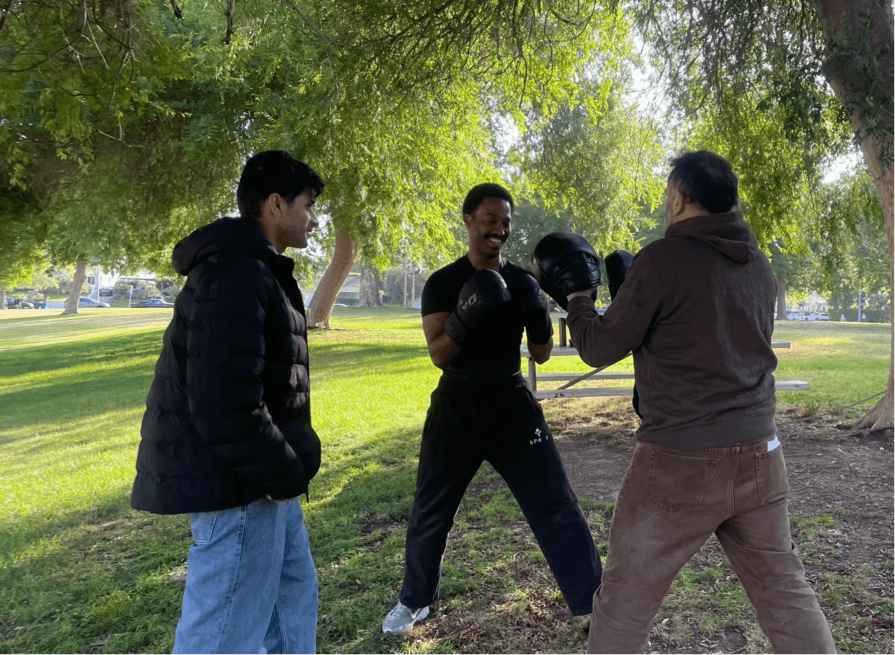 Three people practicing boxing outdoors under trees, with a person in a brown hoodie and gloves, and two others watching.
