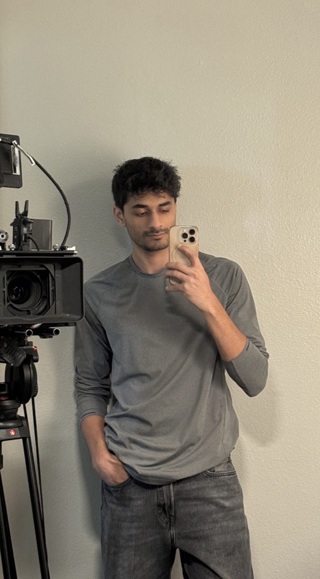 Young man with dark hair and beard taking a selfie in front of a mirror, wearing a gray long-sleeve shirt and jeans.