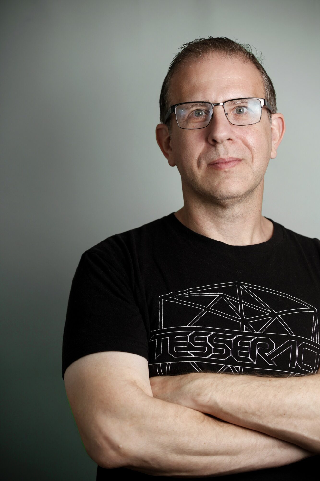 Man with glasses and short hair wearing a black T-shirt with geometric design, arms crossed, neutral expression.