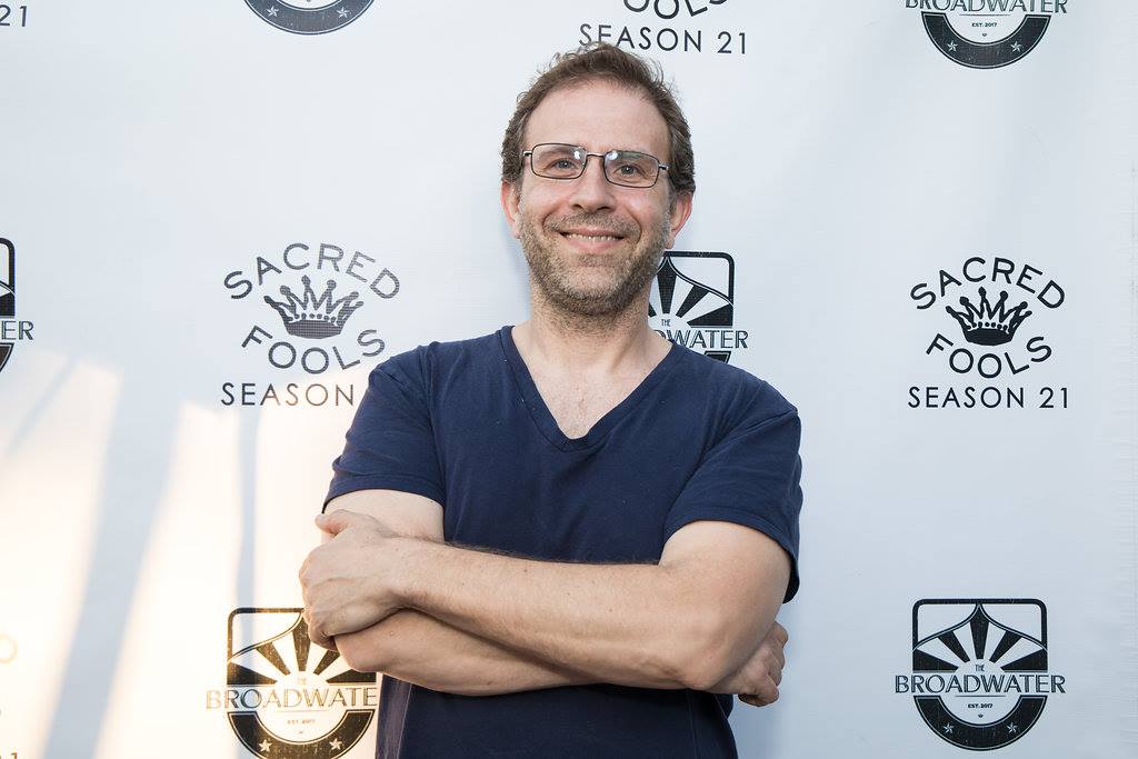 Man with glasses and beard smiling, arms crossed, standing in front of a backdrop with logos and text.