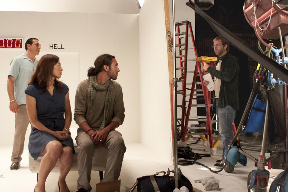 Four people in a studio, two seated and two standing, with a crew member working on equipment in the background.