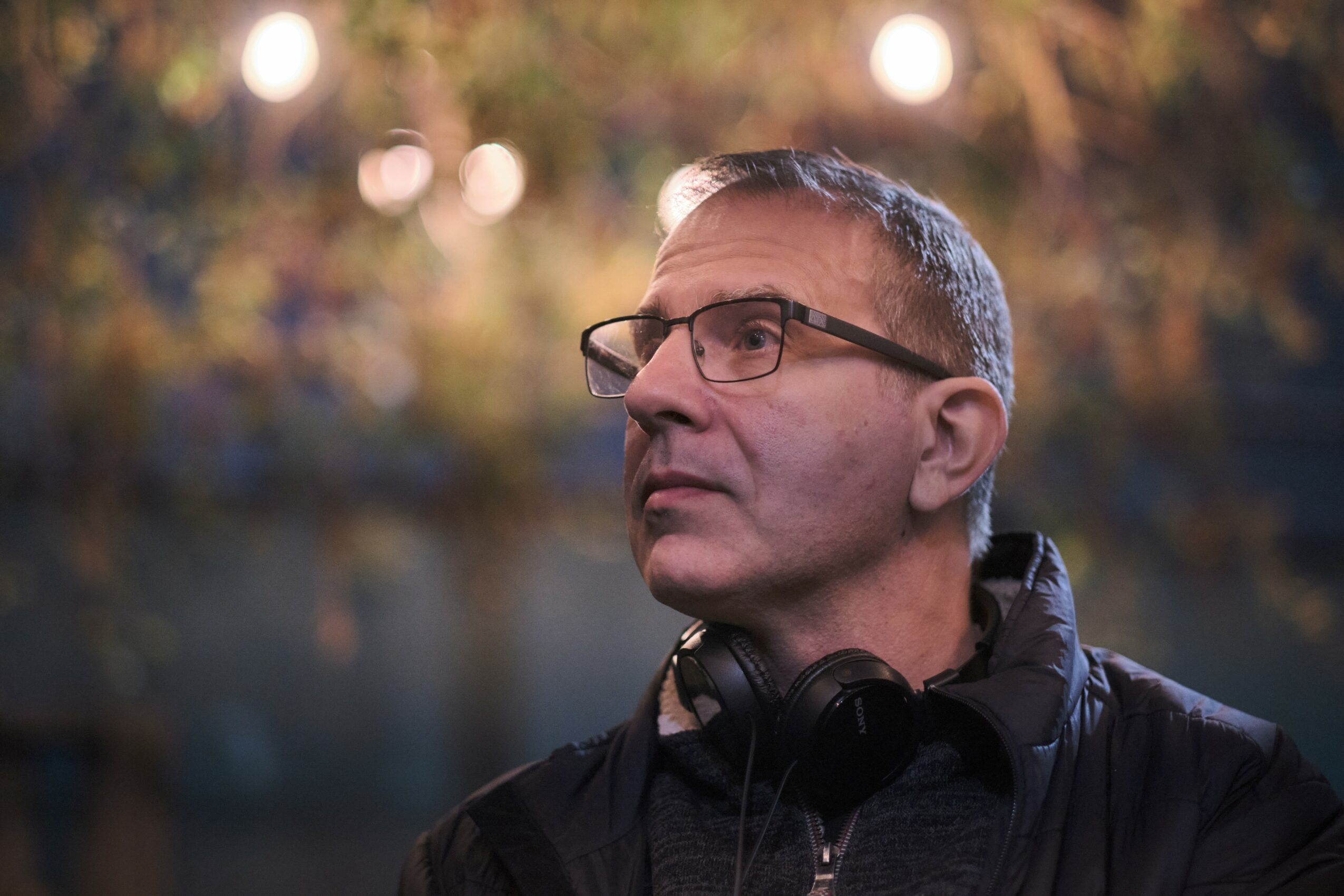 Man with glasses and headphones around neck outdoors, trees in background, looking to the side.