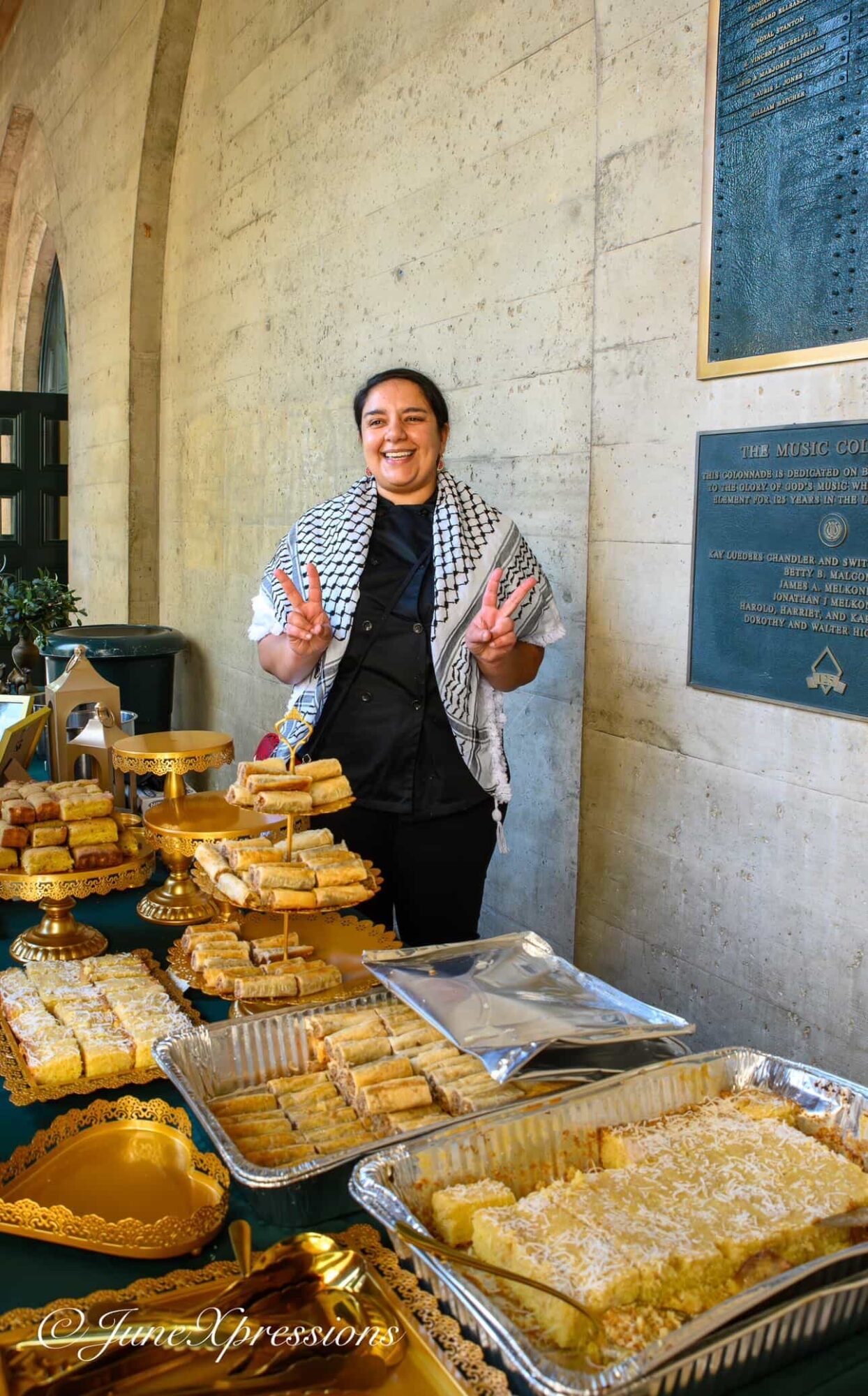 Smiling woman standing behind a table with stacked baked goods, holding a peace sign with both hands, in a stone-walled room.