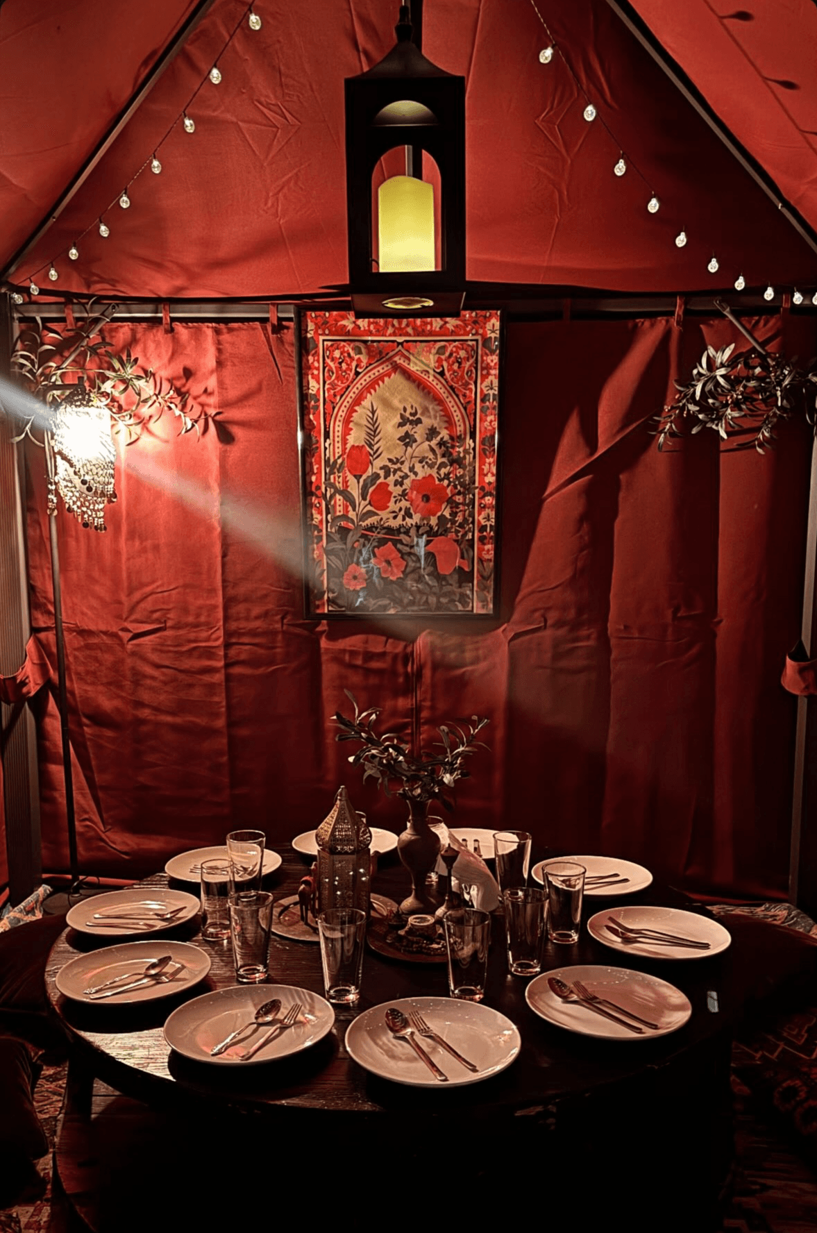 Round table set with plates, glasses, and a floral centerpiece inside a red tent with hanging lights and a lantern.
