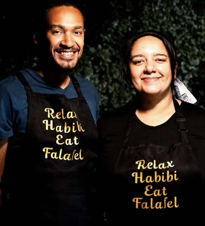 Two people smiling outdoors, wearing black aprons with yellow text, standing in front of greenery.
