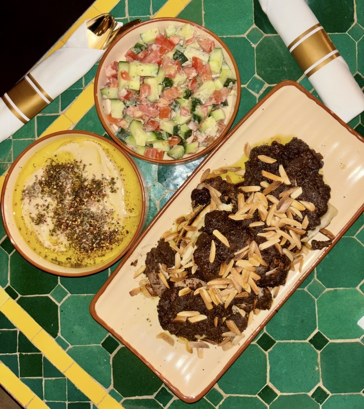 Plate with dark dessert topped with slivered almonds, two bowls with salad and dip, green tiled surface background.