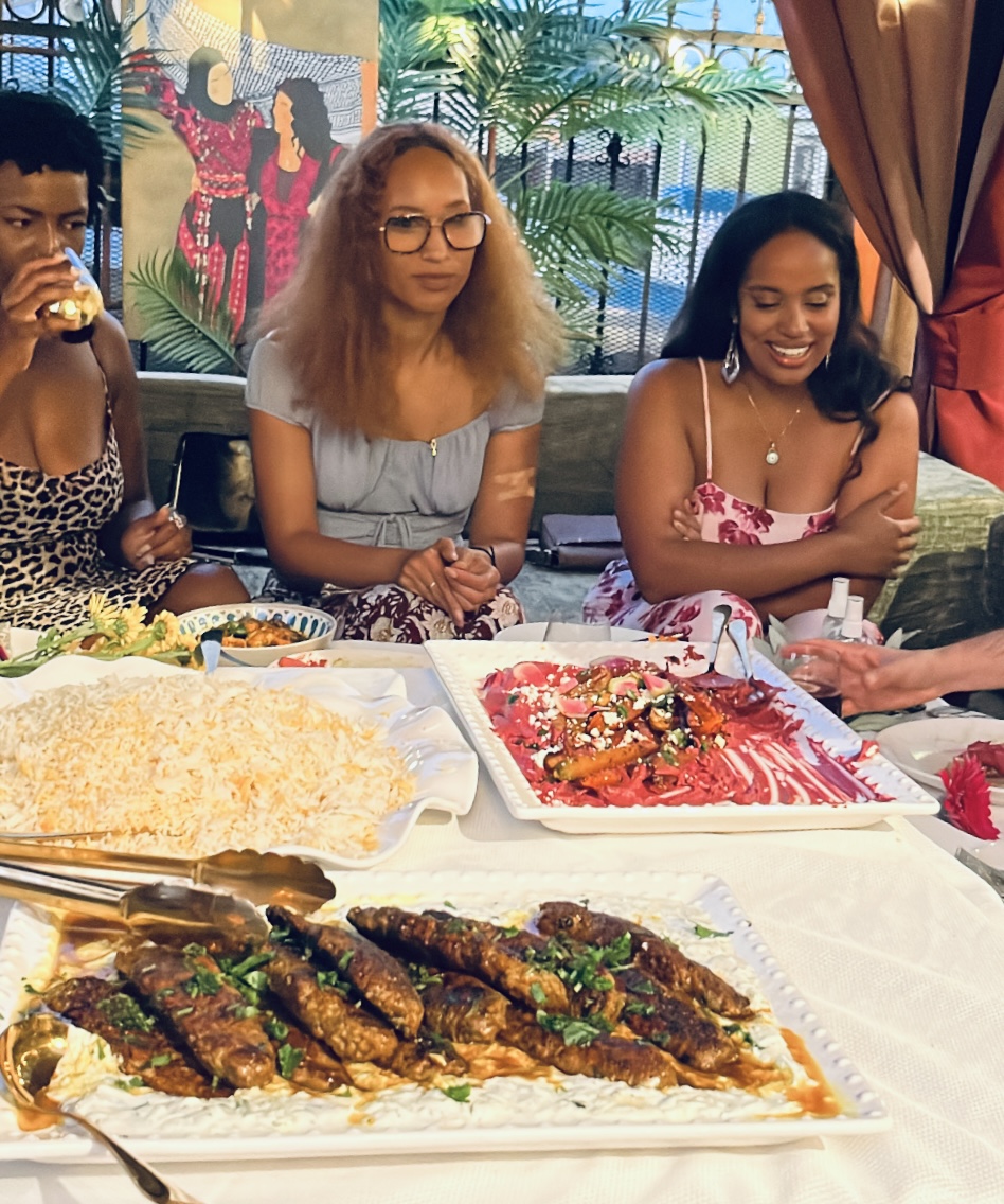 Three women sit at a table with food, including a large cake and grilled dishes, smiling and enjoying the gathering.