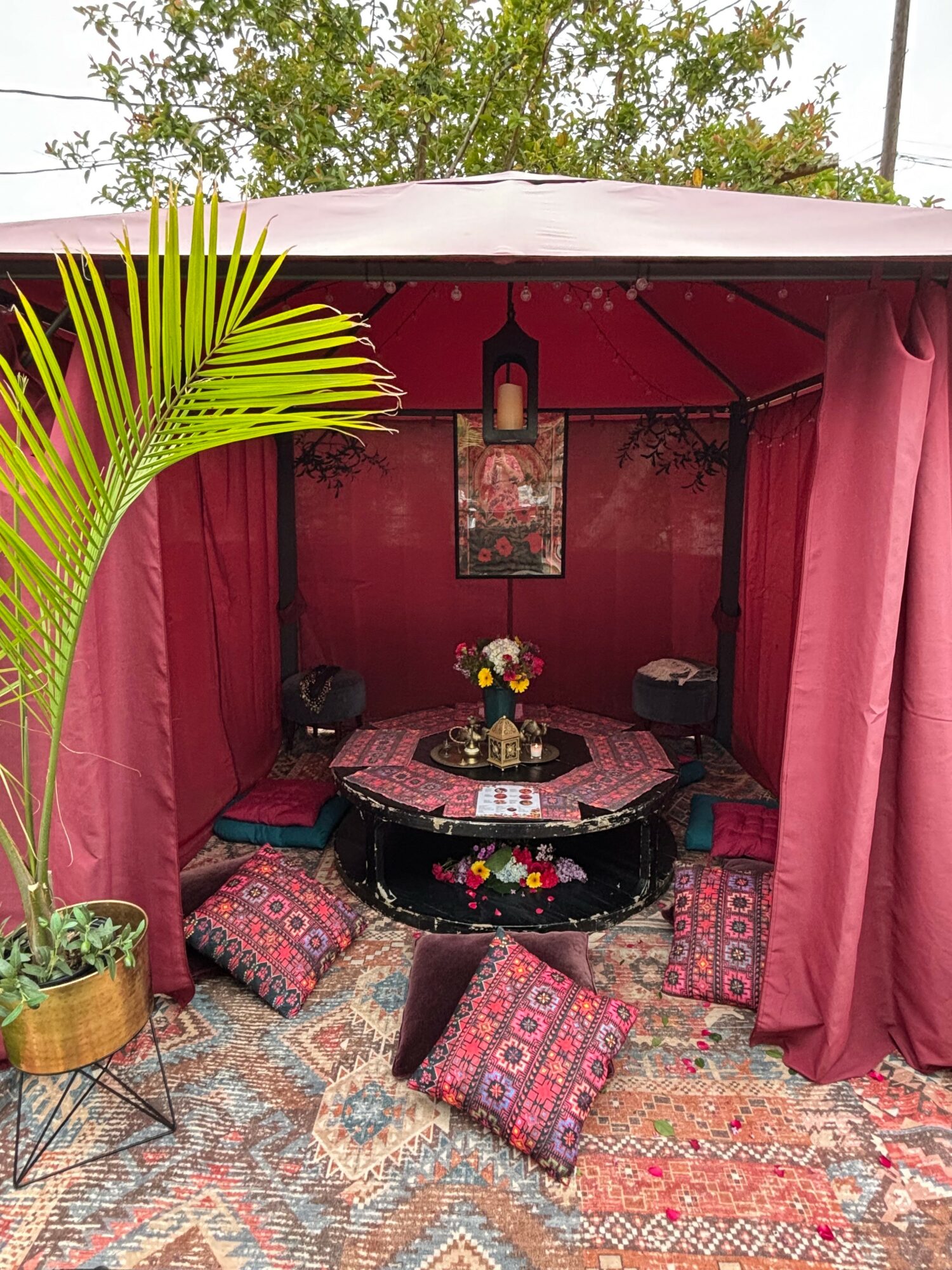 Booth with pink curtains, a round table with a floral centerpiece, cushions on a patterned rug, and a hanging lantern.