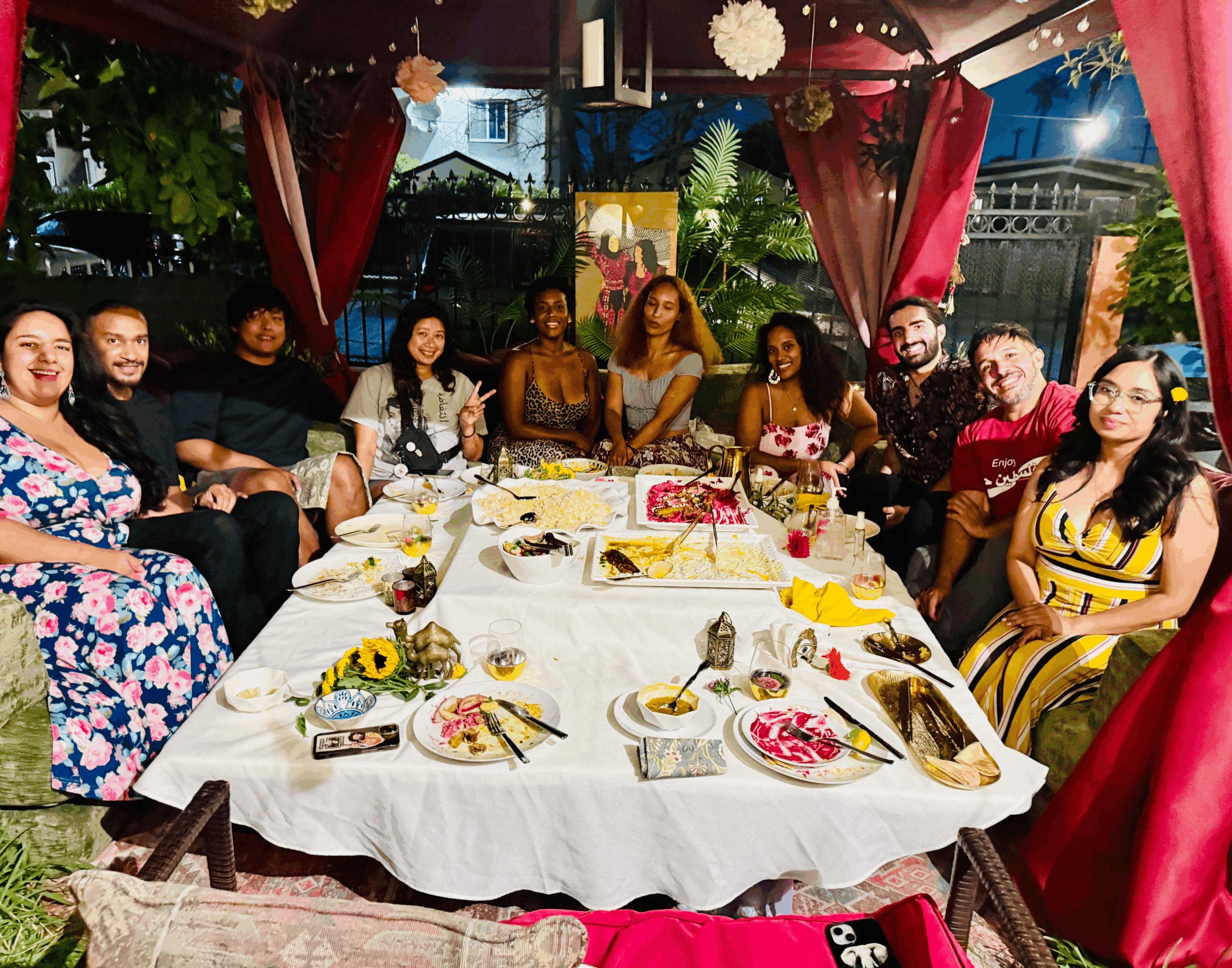 Group of people sitting around a decorated table at a celebration, with colorful curtains and greenery outside.