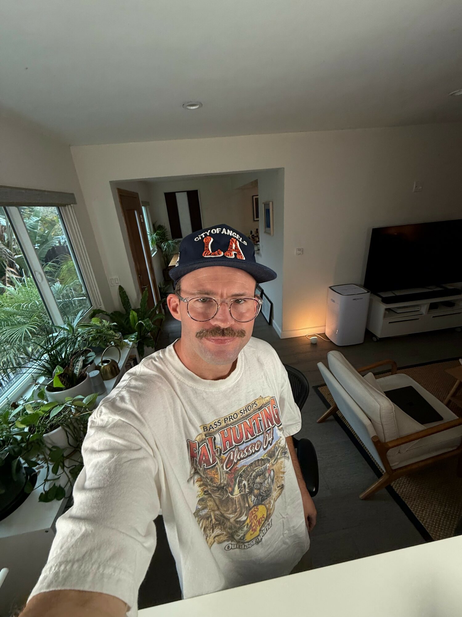 Man wearing a cap and glasses taking a selfie in a living room with plants and furniture