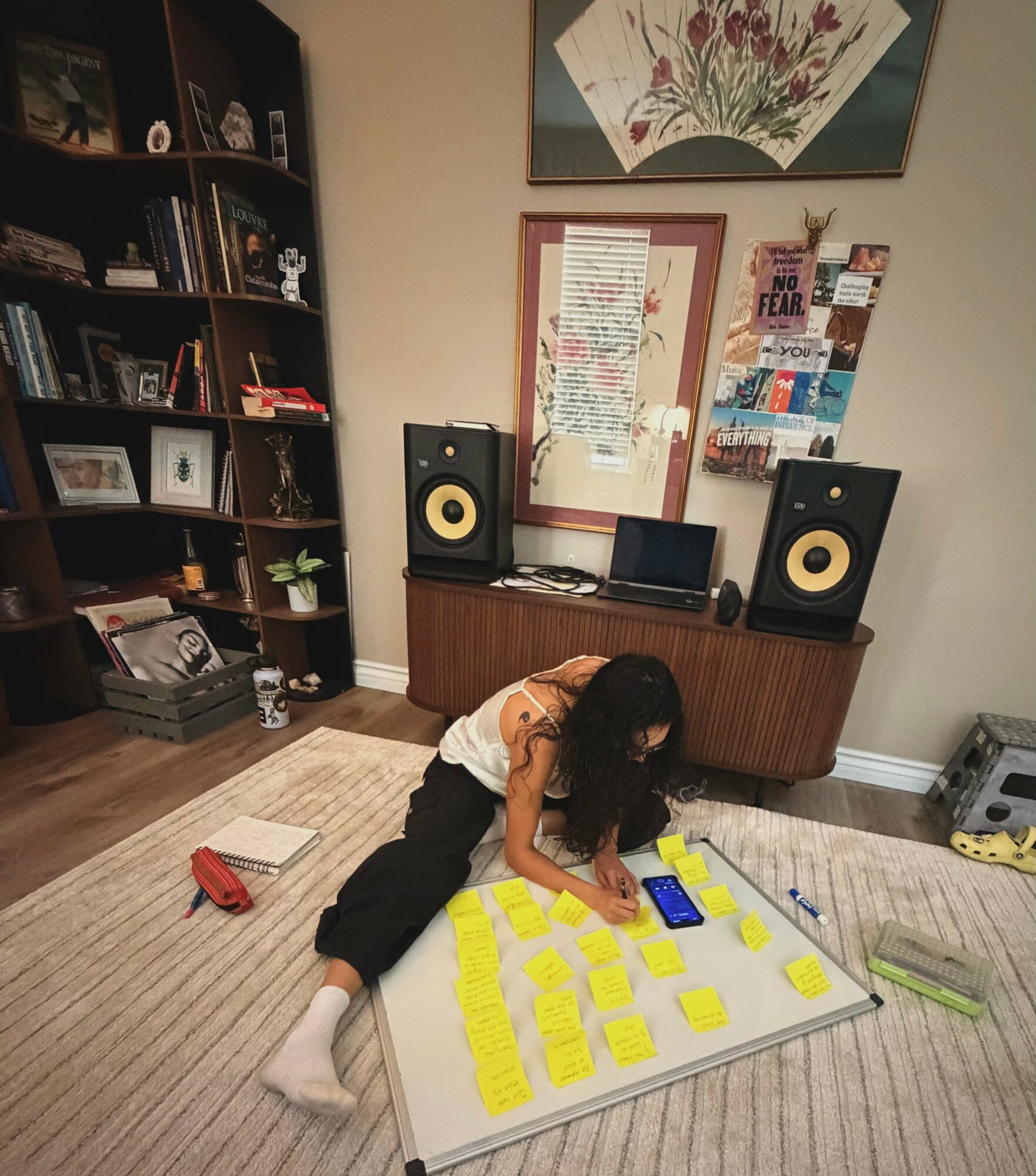 Child with curly hair sits on the floor, writing on a large whiteboard with yellow sticky notes, in a room with bookshelves and speakers.