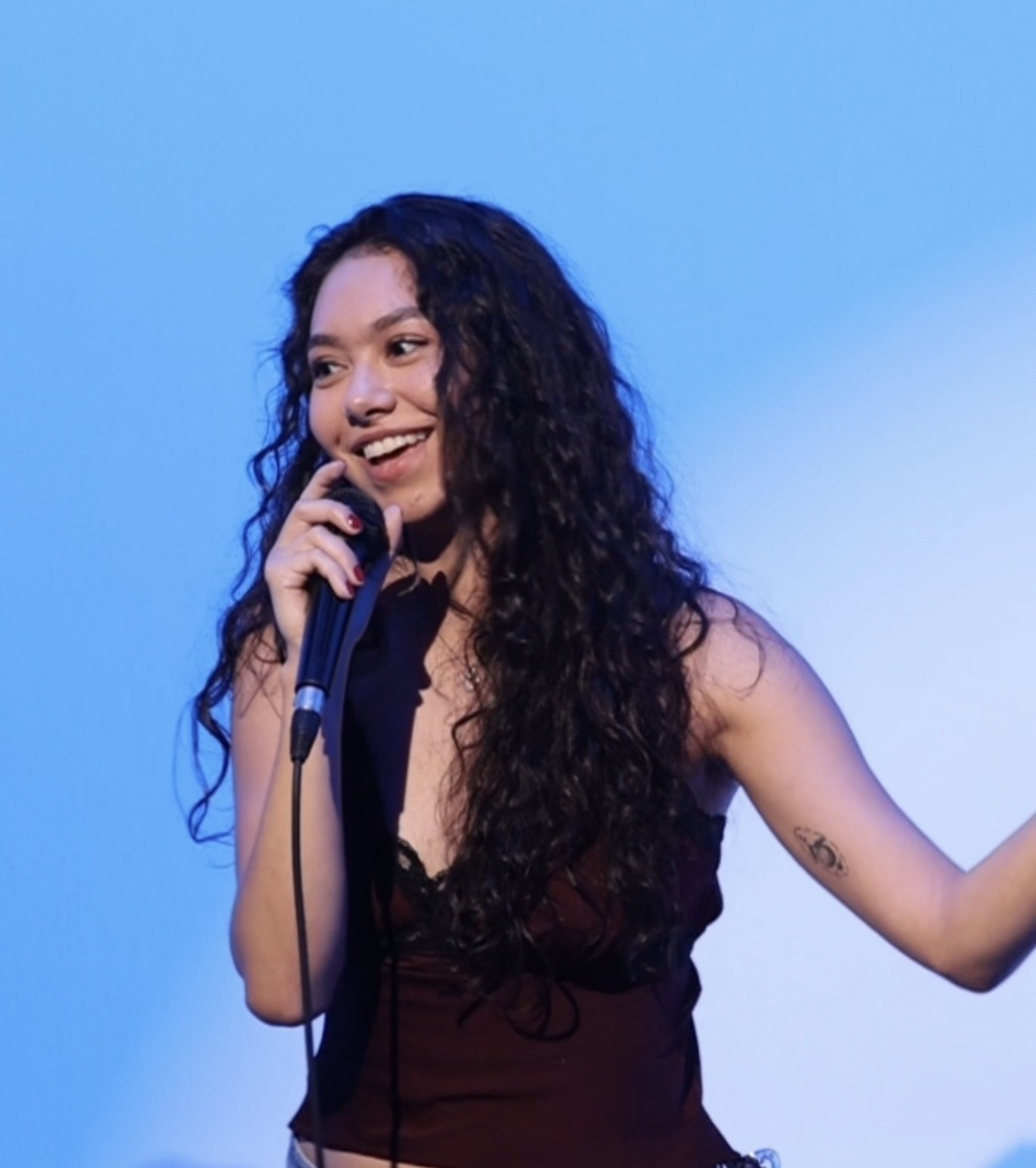 Young woman with long curly hair smiling, holding a microphone, against a blue background.