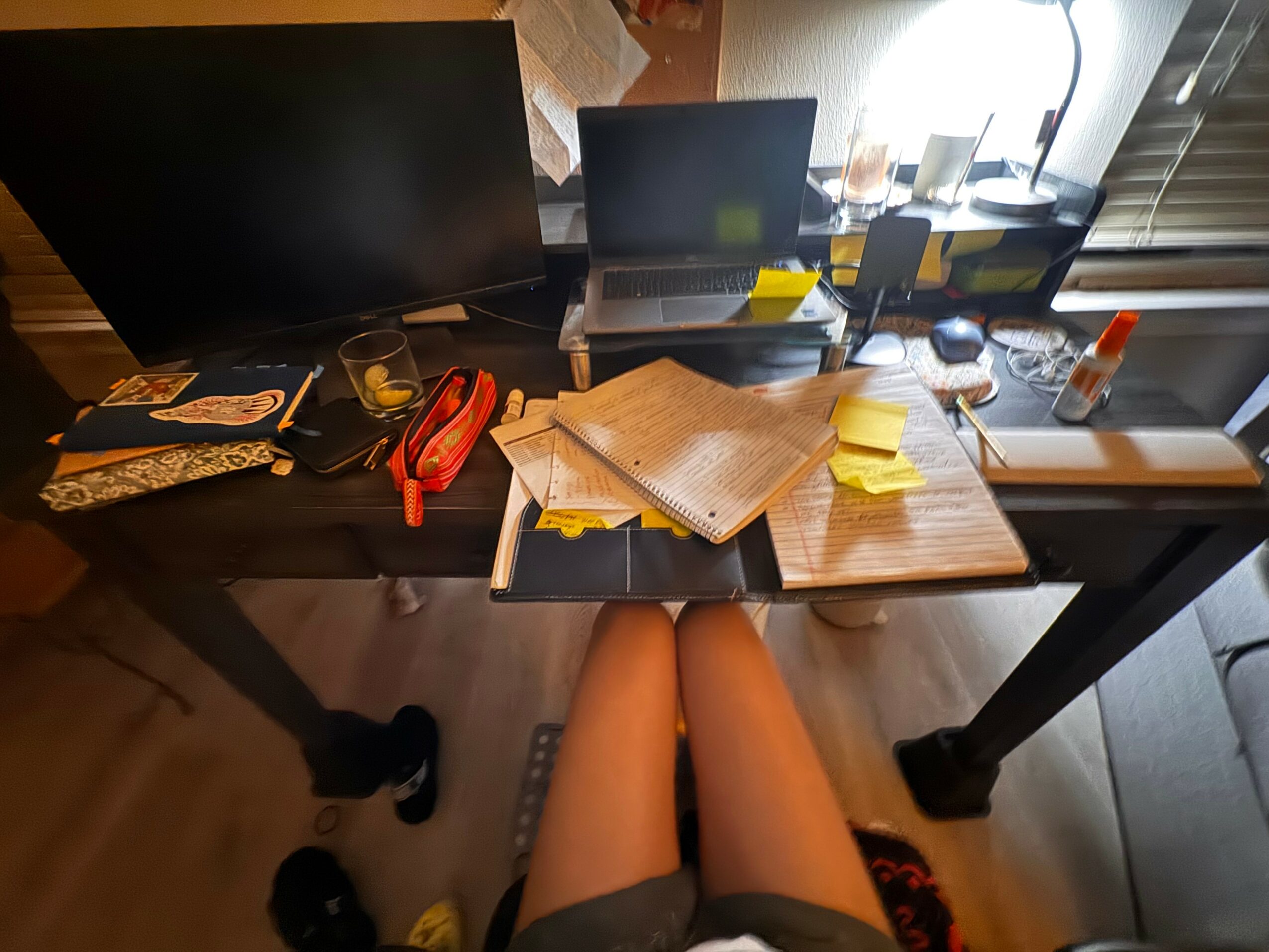 Person sitting at a cluttered desk with papers, notebooks, and office supplies, viewed from above, legs visible.