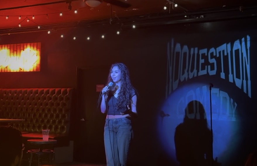 A woman with curly hair stands on stage holding a microphone, with a sign reading 'QUESTION' behind her.