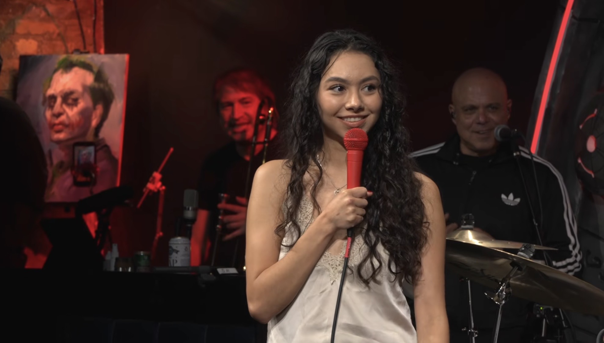 Young woman with long curly hair holding a red microphone on stage, with musicians and a painting in the background.