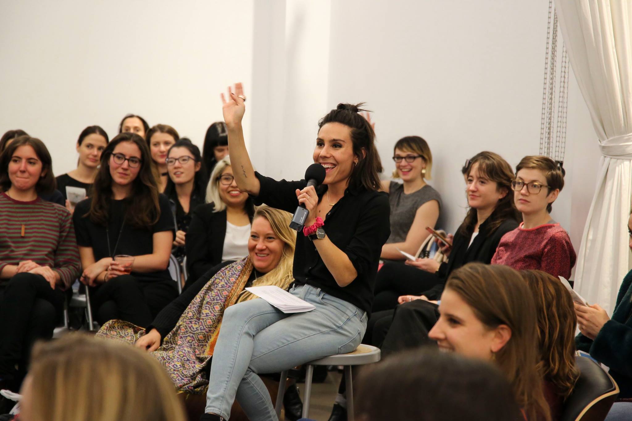 Woman with dark hair, black top, and jeans speaking into a microphone, raising her hand, surrounded by seated audience members.