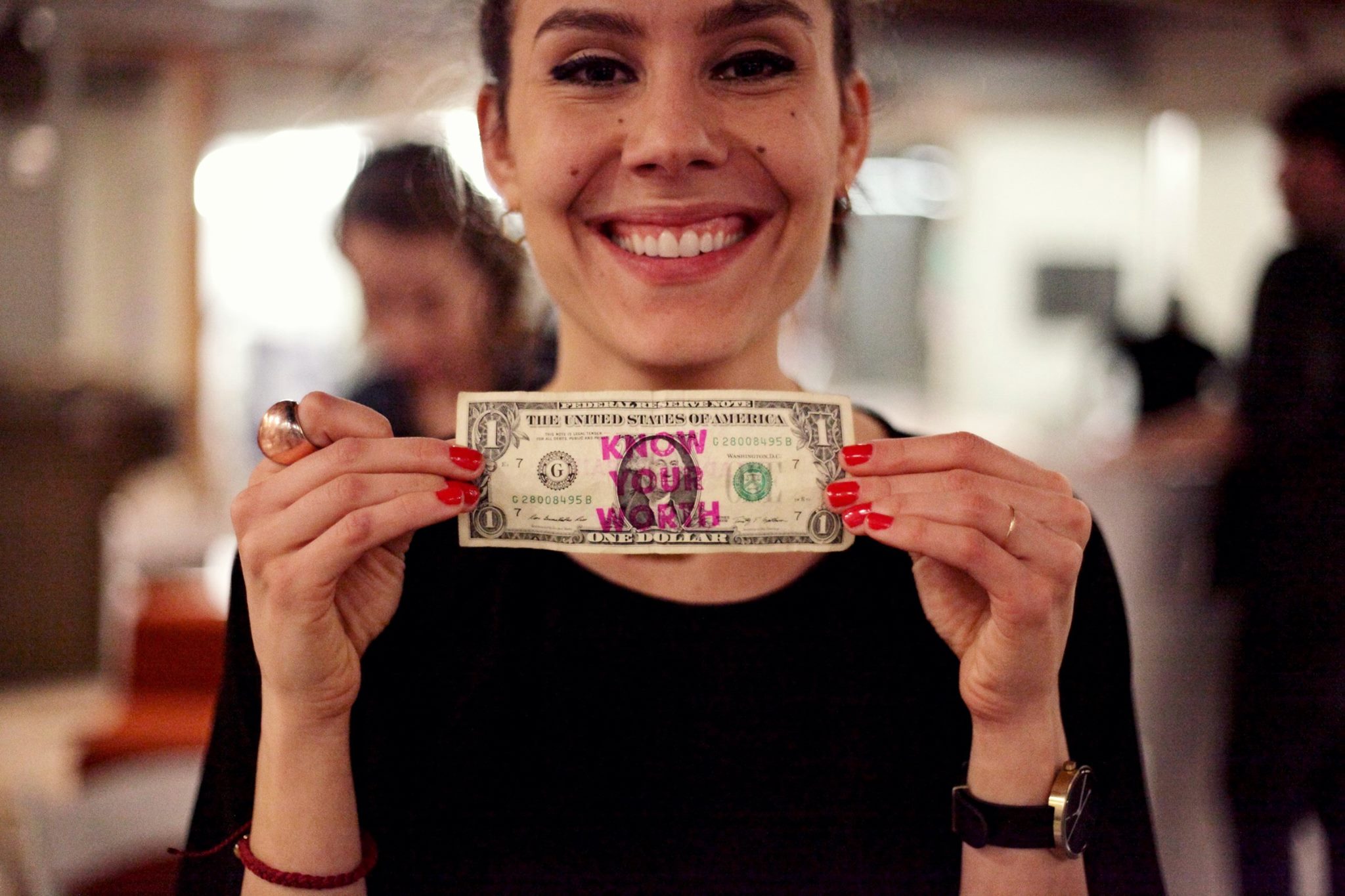 Smiling woman holding a dollar bill with pink text, in a busy indoor setting.