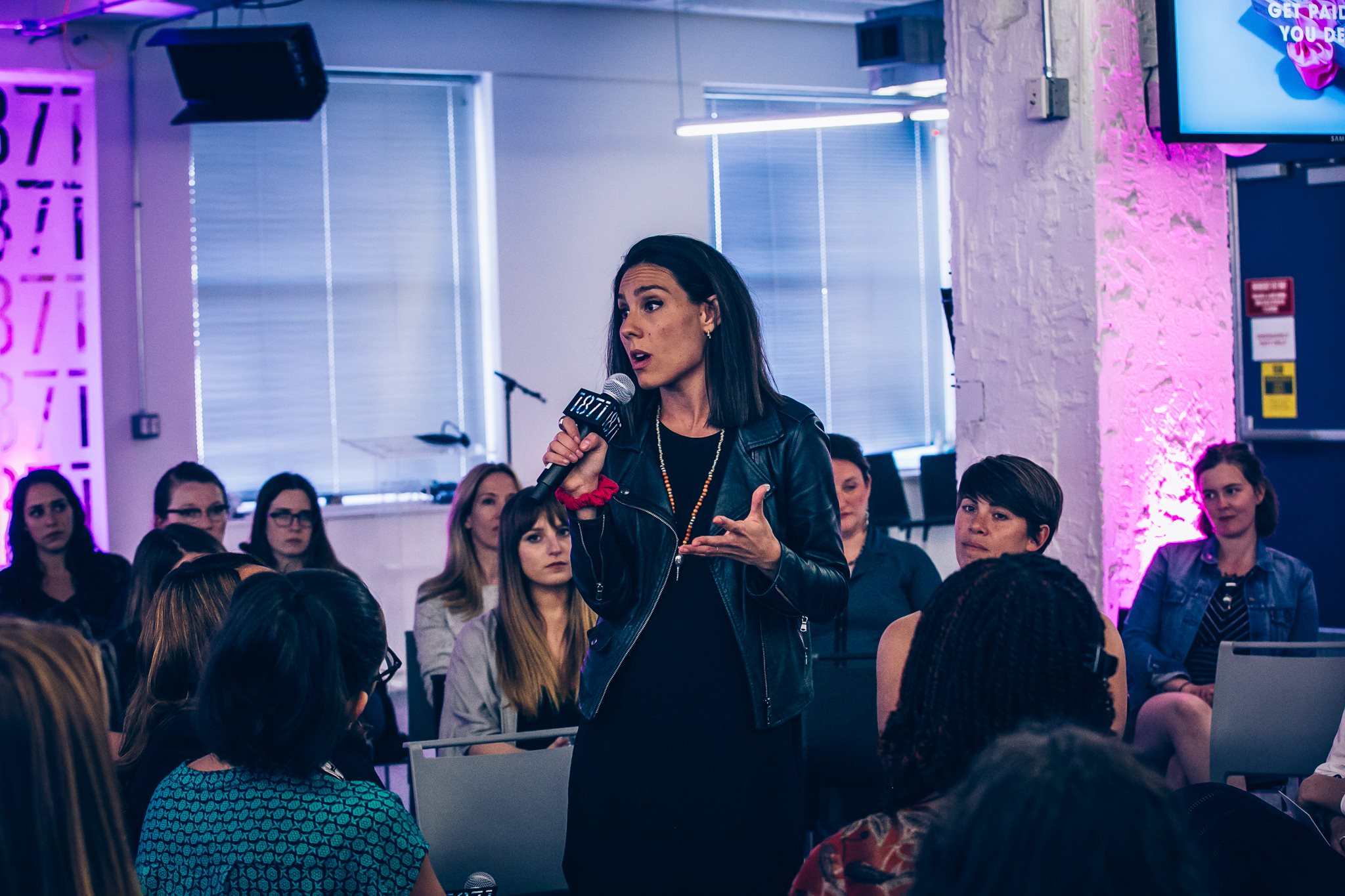 Woman speaking into microphone in front of seated audience in a brightly lit room with pink and purple lighting.