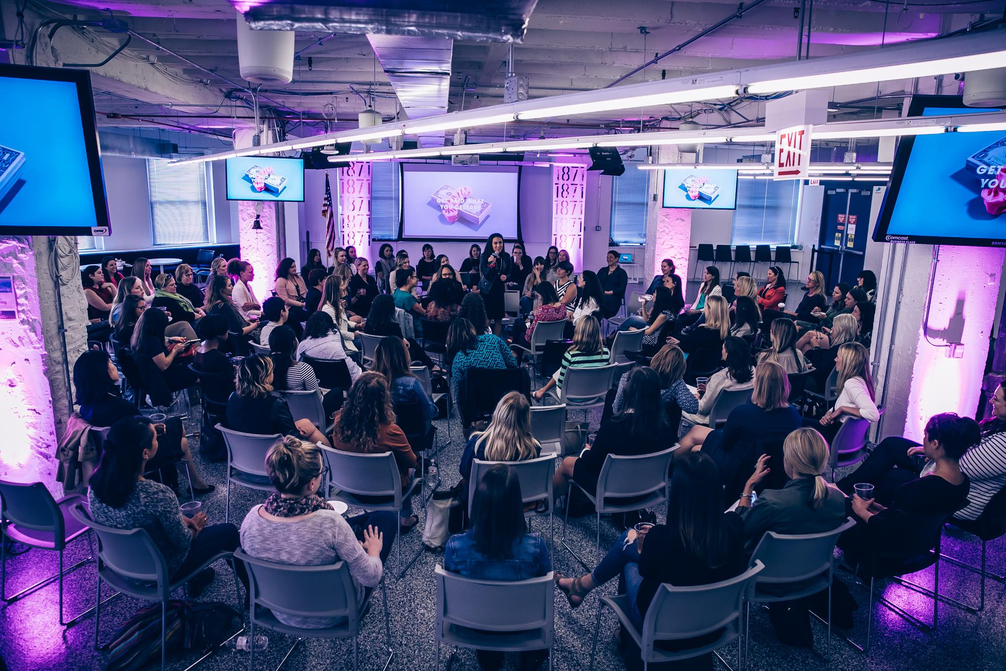 People seated in a conference room with screens and purple lighting, attending a presentation or event.