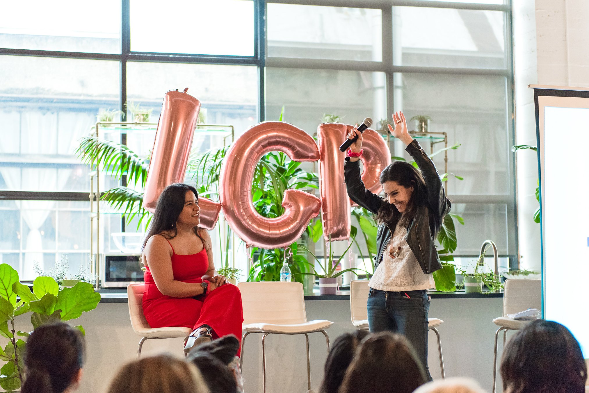 Two women on stage with balloons spelling 'ICN', one sitting and one standing, audience in foreground, large windows behind.