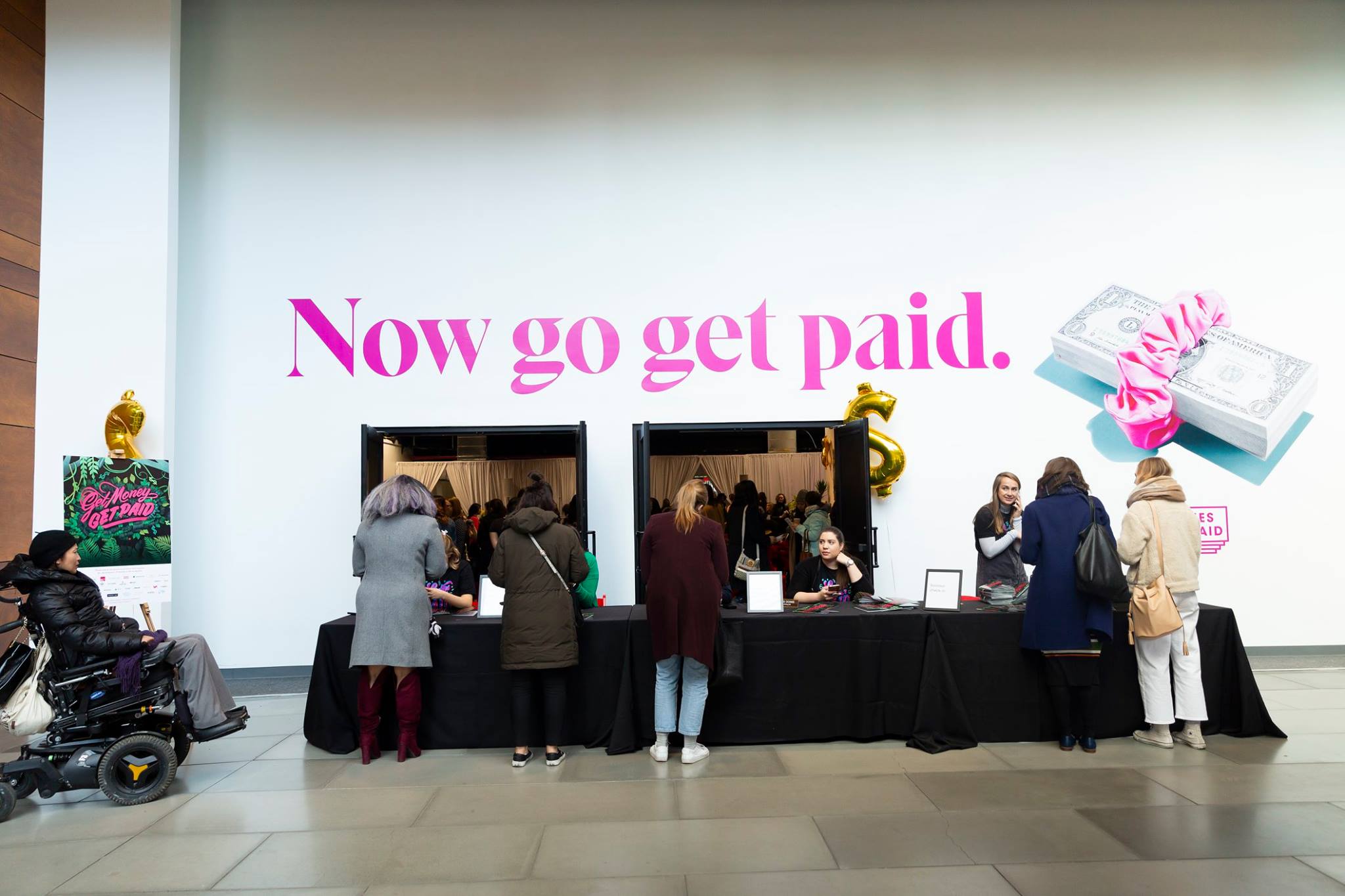 People line up at a payment booth with a large screen displaying pink text and a gift box image.