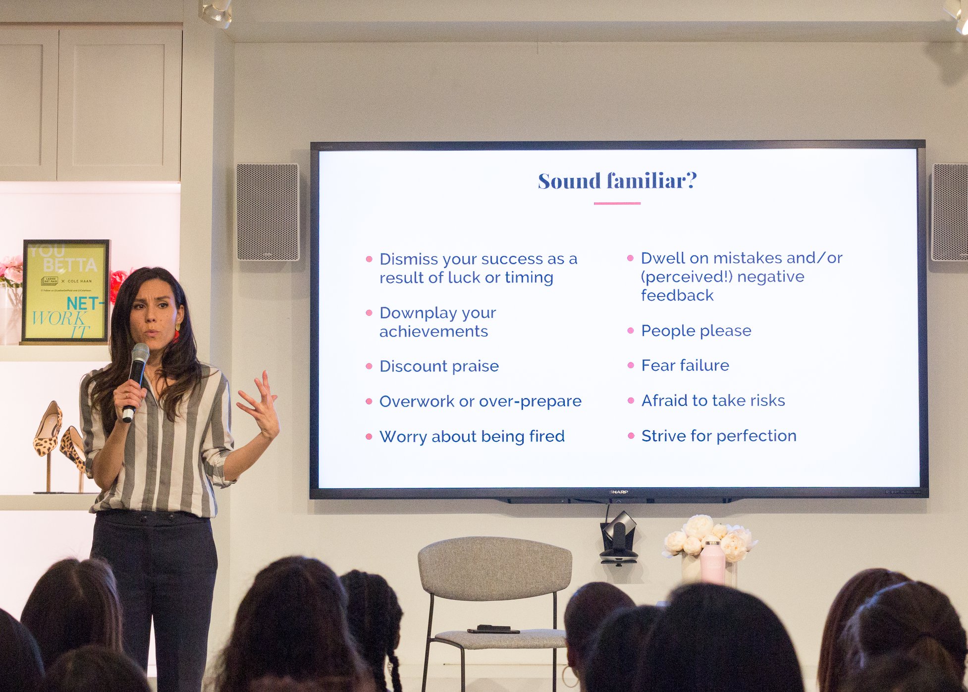 Woman presenting in front of a screen with presentation slide, audience seated, room with white walls and furniture.