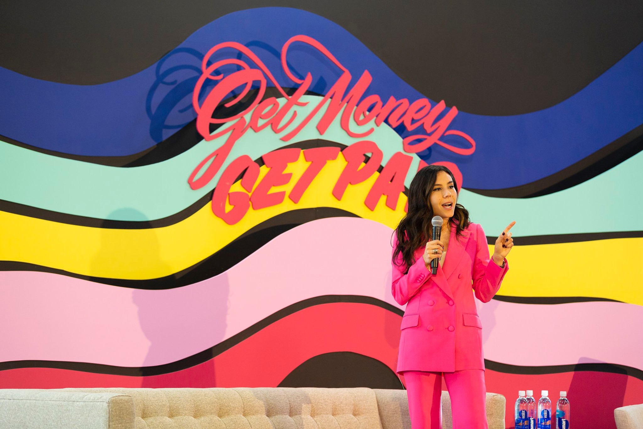 Woman in pink suit speaking into a microphone on stage with colorful abstract background.